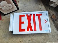 Photo of the rectangular white exit sign light box with red letters and arrow, placed on a concrete floor.
