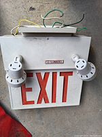 Front view of white Lumacell exit sign featuring large red 'EXIT' letters, fitted with two white emergency lights on each side and exposed electrical wiring on top.