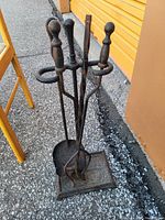 Black metal fireplace tool set including poker, brush, shovel on a black metal stand with decorative base, placed outdoors.