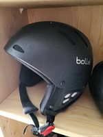 Black Bollé helmet showing side profile on wooden shelf, highlighting ventilation vents, padded lining and chin strap.