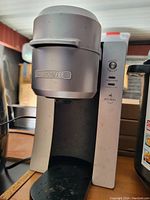 Front angle view of a Keurig coffee maker with Mr. Coffee branding, showing the main body and control panel, placed on a wooden surface with a background of boxes and kitchen appliances.