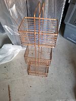 Photo of a three-tier metal wire basket stand with three square wire baskets stacked vertically in a single frame. The basket stand is copper or bronze colored and is resting on a concrete floor with other storage items nearby.