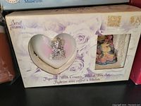 Photo of the Secret Treasures heart-shaped ceramic trinket box and small cherub figurine set inside original box showing floral theme and design details.
