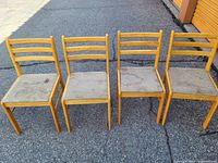 Four wooden ladderback chairs with beige fabric seats, placed side by side on an asphalt surface. The chairs show visible wear and staining on the seats.