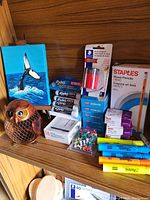 Image of various office supplies arranged on a wood shelf including markers, pencils, tapes, push pins, a decorative owl figurine, and a small orca canvas art.