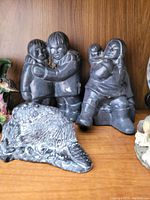 Three dark colored figurines/statues arranged on shelf: two children figurines and one animal statue.