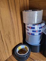 Photo depicting four rolls of tape stacked vertically against a wooden panel background, includes clear packing tape, branded packaging tape, silver duct tape, and black tape.