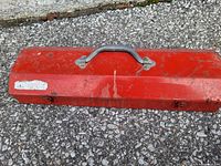Closed red metal toolbox resting on asphalt surface, showing exterior with carry handle and latch locks.