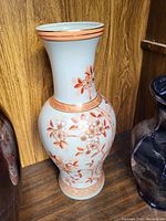 Photo of tall white porcelain vase with orange floral designs sitting on wooden shelf.