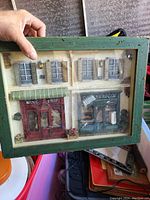 Hand holding a framed wall decor piece depicting two vintage shops: a red Wine Shop and a green Grocer with detailed 3D architectural elements.