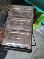 Electric warming tray with three stainless steel pans lined up, each having a transparent plastic lid with a handle on top. The tray has a black base with an electric cord visible.