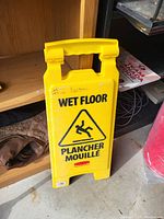 Yellow plastic wet floor sign standing upright on a shelf. Visible bilingual text and warning symbol.