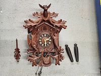 Front view of the assembled antique wooden cuckoo clock with deer head, fish, oak leaf carvings, clock face, and two pine cone weights attached by chains laid out.
