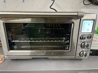 Front view of the Breville toaster oven showing stainless steel body, glass door, and control panel with knobs and LCD screen.