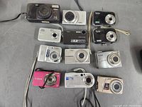 Top view of twelve assorted old digital cameras showing fronts with lenses and a few wrist straps.