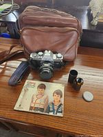 Photo of Canon AL-1 camera with lens, manual, small film canister, and brown leather carrying case on wooden surface.