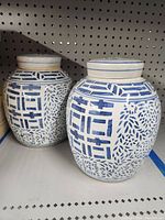 Pair of white ceramic urns with blue geometric and leaf brush painted decoration, displayed side by side on shelf.