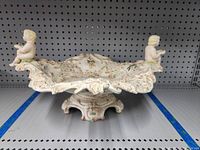 Front view of the ornate decorative porcelain bowl with two cherubs on the rim showing detailed floral patterns and scalloped edges.