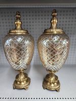 Pair of clear glass jars with diamond cut lattice pattern and gold-tone finials and bases on white pegboard shelf.