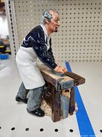 Side view of porcelain carpenter figurine wearing blue shirt, white apron, gray pants, working on a wooden workbench with a hand plane tool.