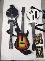 Three Guitar Hero guitars (two for Wii, one for Xbox 360) arranged flat with straps and control buttons visible alongside two packs of stickers.