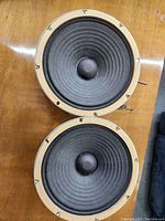 Photo showing two mid-sized Jensen speaker cones mounted on a wooden surface, cones black with visible dust caps and mounting holes on metal frames.