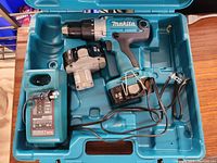 Open teal plastic tool case showing cordless Makita power drill, two batteries, and battery charger coiled inside