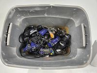 Overhead view of a gray plastic bin filled with approximately 11 assorted wired video game controllers in various colors and conditions, cords tangled.