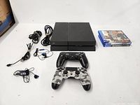 Full lot shot showing PS4 console, 2 controllers (black and white camo), 3 PS4 games, 2 charging cords, HDMI cable, power cord, and 2 earbud headsets arranged on white surface.