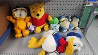 Photo of the collection of plush dolls including Garfield, Winnie the Pooh, Bulbasaur, Snorlax, and Donald Duck on a pegboard shelf.