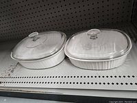 Two oval Pyrex glass baking dishes with matching clear lids. The dishes have ribbed sides and are opaque white on the outside with clear glass lids featuring the Pyrex logo.