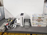 Nintendo Wii console with missing GameCube controller port cover, stack of 10 Wii game cases to the right, various cables and controllers to the left.