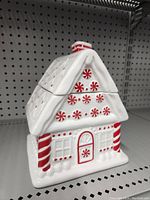 Front view of white ceramic gingerbread house cookie jar with red and white peppermint decorations and candy cane pillars on store shelf.