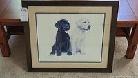 Front view of framed artwork showing two Labrador puppies, black and yellow, signed and numbered in lower left corner, dark wood frame and double matting
