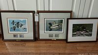 Framed prints with signatures and duck stamps displayed beneath each print, showing mallards and flying waterfowl.