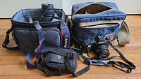 Two Canon cameras (T50 and Rebel 2000) with lenses, instruction manual, and two carry cases on wooden floor. Both cameras have straps. One carry case holds a lens and camera items.