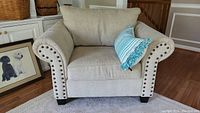 Front view of beige fabric armchair with studded arms and decorative pillow on seat.