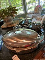 Cookware set of two pots with glass lids and one covered roasting pan on kitchen counter near window.
