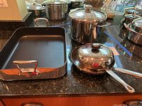 Three Kitchenaid stainless steel cookware pieces: one tall stockpot with lid, one saucepan with lid, one sauté pan with lid, and a rectangular Mauviel roasting pan with non-stick interior and polished metal handles.
