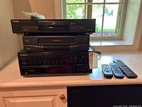 Three stacked black electronics on a white counter near a window: Toshiba DVD player SD-2109 on top, Magnavox VHS player under it, and Pioneer stereo receiver VSX-455 at the bottom. Three remote controls are placed on the counter beside them.