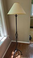 Full view of the floor lamp showing the tripod base, decorative twisted metal stem, and beige fabric shade leaning slightly next to a window.
