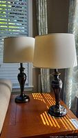 Two matching table lamps with dark bronze metal bases and cream-colored fabric drum shades placed on wooden table near window. Visible dent on the base of lamps.