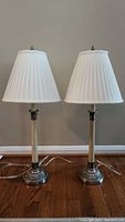 Pair of matching table lamps with white pleated lampshades and distressed stands on a wood floor against a beige wall.