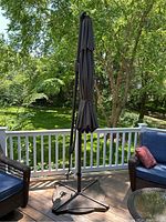 Umbrella closed and standing next to outdoor chairs on a deck with white railing and green trees in background.