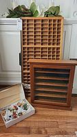 Large wooden printers drawer standing upright with many small square compartments visible; next to it, a smaller wooden golf ball display case with glass front and horizontal shelves; box of assorted miniatures on floor in front.