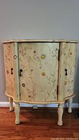 Front view of cream distressed side table with floral painted doors and black metal hardware on four legs.