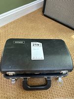 Closed black Yamaha branded hard case on carpet floor with a Fender amplifier in background.