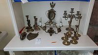 Full lot view of brass door knocker, candlestick holders, shield plaque, and wall light fixture arranged on white shelf