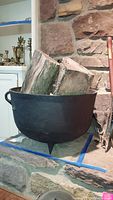 Front view of vintage cast iron cauldron holding several large pieces of firewood, placed on a stone hearth in front of a stone wall.