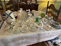 Wide view of table showing blue and white windmill cookie jar, porcelain cups and saucers with floral designs, various glassware including swan servers, pewter plates, and silverware box.